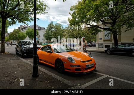 Vibrant orange Porsche 911 GT3 RS parked in a designated spot, surrounded by lush trees and bustling with the energy of swiftly passing cyclists. Stock Photo