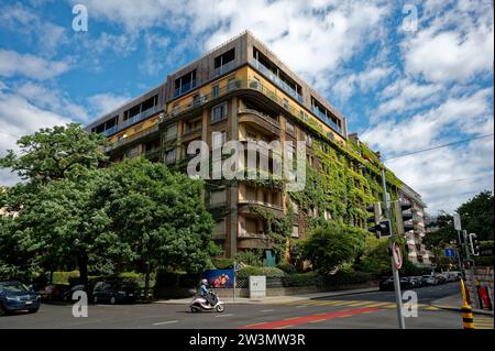 An imposing residential building adorned with lush, bright green foliage on its exterior, nestled in a charming corner of Geneva as a scooter passes. Stock Photo