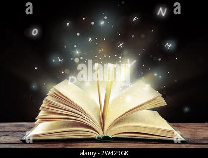 Open book with magic light and glowing letters flying out of it on wooden table against black background Stock Photo