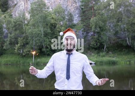 Smiling bearded guy or man in santa hat holds a sparkler in his hands and wishes a happy new year. Against the background of the summer landscape. Stock Photo
