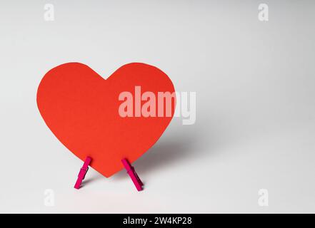 Red paper heart stands on wooden clothespins on white background. Minimal love concept. Space for text. Stock Photo