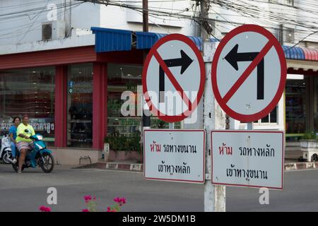 a road junction in thailand with road signs indicating no left and no right turns, with written confirmation, as a motorcyclist and passenger look on Stock Photo