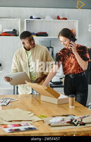 smiling asian woman talking on smartphone near african american man with laptop and carton box Stock Photo