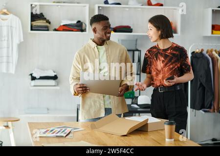 happy interracial designers with gadgets talking near carton box with clothes in print studio Stock Photo