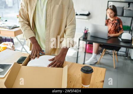 african american man packing clothes in carton box near asian designer at laptop in print studio Stock Photo