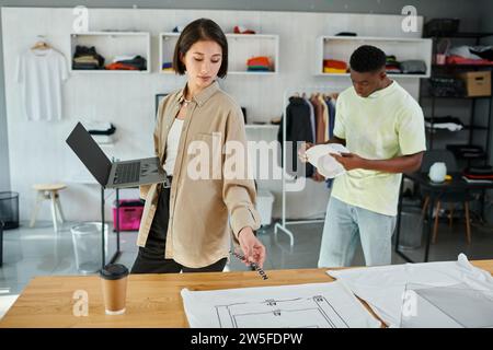 asian designer with laptop and text template near clothes and african american man in print studio Stock Photo