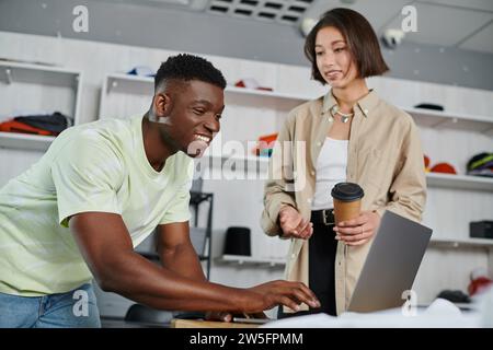 cheerful african american man using laptop near asian colleague with coffee to go in print studio Stock Photo