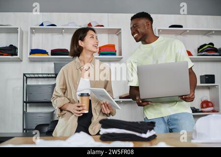 joyful interracial designers with devices and coffee to go talking near clothes in print studio Stock Photo