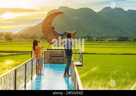 Man using camera to take photo with happiness young couple on crescent moon chair made of rattan in paddy field with beautiful scenic in evening. Deco Stock Photo