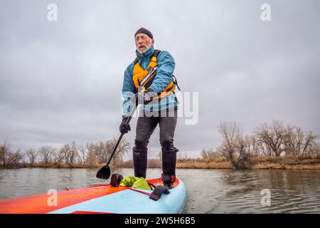 senior male paddler is paddling a stand up paddleboard in cold winter weather on a calm lake Stock Photo