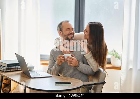 cheerful man with coffee cup sitting near laptop and caring wife in home office, child-free couple Stock Photo