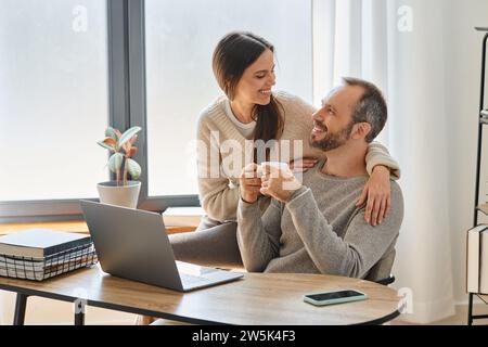 happy man with coffee cup sitting near laptop and caring wife in home office, child-free lifestyle Stock Photo
