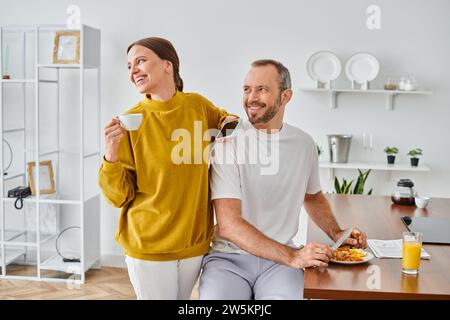 happy man enjoying tasty breakfast near wife with coffee cup and smartphone looking away in kitchen Stock Photo