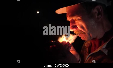 A man lights a cigarette at night on the street in the  dark.Night,darkness,smoking cigarettes and exhaling tobacco smoke.Night  street.The fire from the match illuminates the persons face.Tobacco smoke