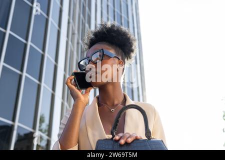 From above of Beautiful African American businesswoman talking on smartphone while sitting with black purse in urban city against building Stock Photo