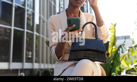 Crop hands of anonymous African American young businesswoman sitting with black purse and using smartphone in city Stock Photo