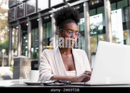 Focused young African American businesswoman with curly hair wearing eyeglasses and working over laptop while sitting at sidewalk cafe Stock Photo