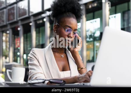 Focused calm young African American businesswoman having business call over mobile phone and using laptop at sidewalk cafe Stock Photo