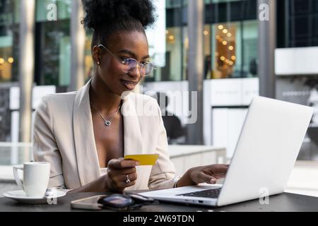 Smiling African American young female executive holding credit card and using laptop for online payment at sidewalk cafe Stock Photo
