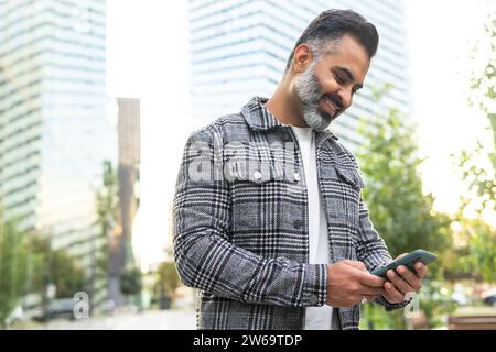 Positive bearded Indian businessman in checkered jacket smiling and browsing smartphone against blurred cityscape background Stock Photo