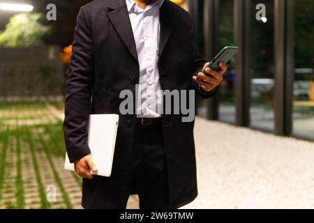 Faceless male entrepreneur in classy outfit holding laptop and messaging via smartphone after successful business meeting Stock Photo