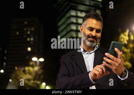 Smiling bearded indian man in suit standing against glowing lamps and buildings at evening time outside while texting with cellphone Stock Photo