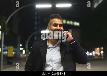 Smiling bearded indian man in suit standing on street with streetlights and talking on cellphone against illuminated city buildings Stock Photo