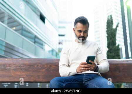 Concentrated bearded indian man in jeans and white sweater sitting alone on street wooden bench while using smartphone against modern buildings of cit Stock Photo