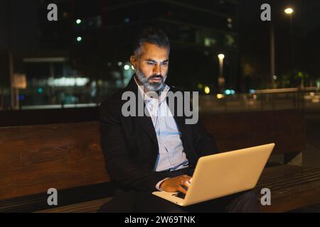 Serious bearded businessman in suit surfing internet with netbook while sitting on wooden bench at evening time against illuminated buildings of moder Stock Photo