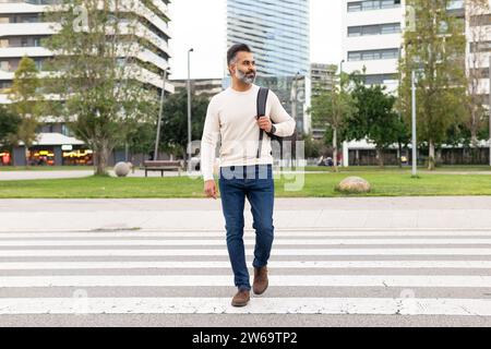 Full body of Bearded adult businessman wearing white sweater with rucksack crossing the street and looking away against background of skyscrapers and Stock Photo