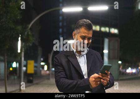Smiling bearded indian man in suit standing on street with streetlights and texting with cellphone against illuminated city buildings Stock Photo