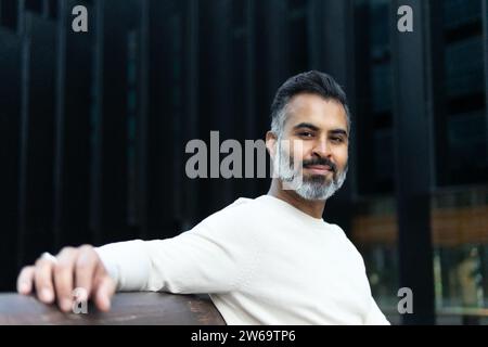 Smiling bearded indian businessman in casual outfit sitting alone and leaning on back of wooden bench while looking at camera Stock Photo