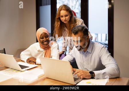 Concentrated multiethnic colleagues in casual clothes sitting at table with laptops and working on business project in modern office Stock Photo