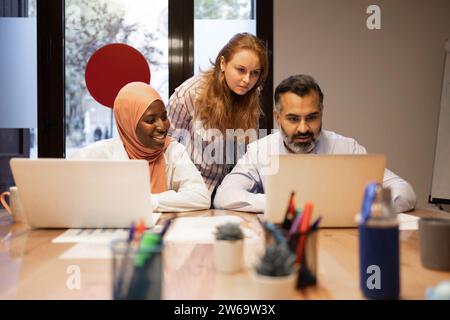 Concentrated multiethnic colleagues in casual clothes sitting at table with laptops and working on business project in modern office Stock Photo