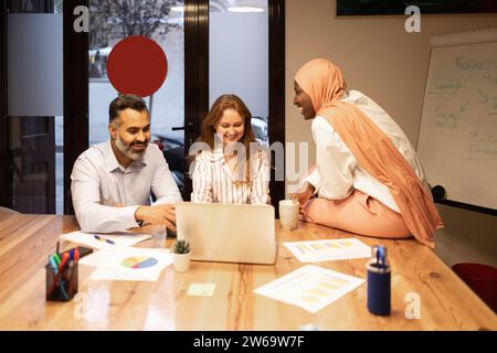 Smiling diverse coworkers sitting at table with papers and laptop while discussing business plan in modern office Stock Photo