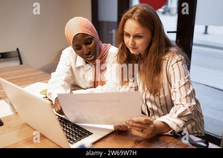 Positive multiethnic young female colleagues sitting at desk using laptop and working on project in modern office while discussing business strategy Stock Photo