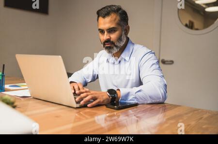 Focused bearded male manager sitting at wooden table and using laptop to work on business project in in modern office Stock Photo