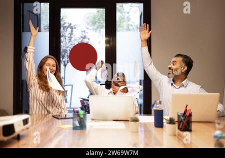 Happy young diverse colleagues raising arms and celebrating success while sitting at table with laptops in modern office Stock Photo