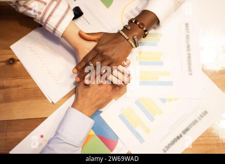 Top view of crop diverse colleagues stacking hands on wooden table with charts papers in modern office during work process at desk Stock Photo