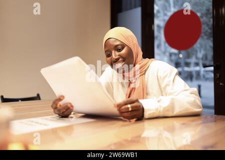 Smiling muslim businesswoman wearing headscarf sitting at table with papers and reading documents while working on creative project in modern office Stock Photo