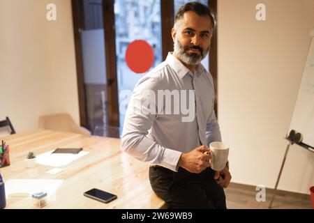Happy bearded businessman with coffee cup sitting at wooden table and looking at camera in modern office against blurred background Stock Photo