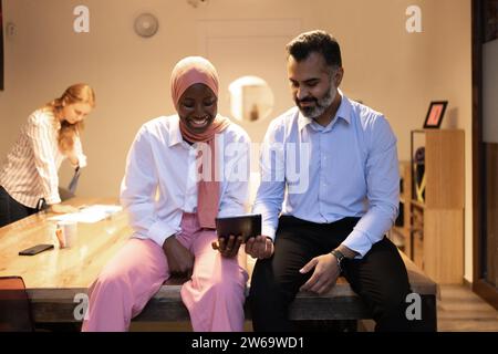 Smiling diverse colleagues sitting at wooden table and browsing tablet while discussing project details in modern office with blurred background of fe Stock Photo
