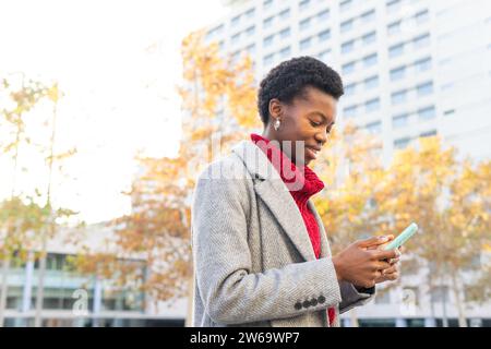 Smiling African American female browsing mobile phone while standing against blurred modern buildings in city street Stock Photo