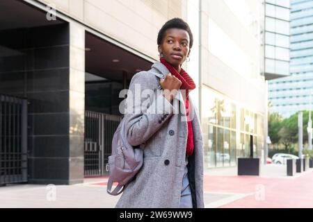 Confident African American woman wearing gray coat and looking away while standing on street near modern buildings Stock Photo