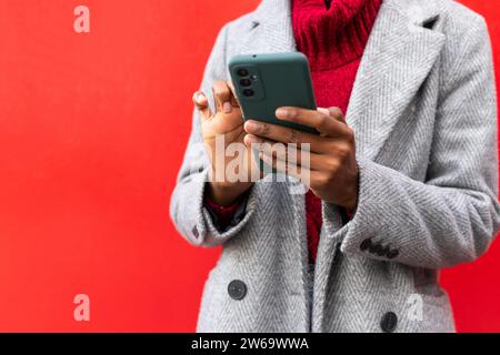 Cropped unrecognizable African American lady in jacket browsing smartphone while standing against red background Stock Photo
