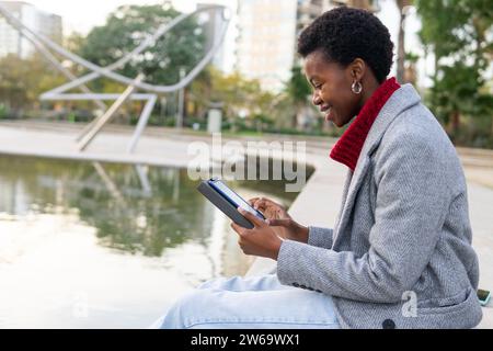 Side view of smiling African American female in outerwear sitting near pond and reading electronic book while resting in park during weekend Stock Photo