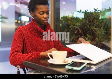 African American female remote worker typing information on netbook while drinking cappuccino in cafeteria Stock Photo