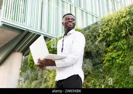 From below of young African American male freelancer in white shirt holding netbook while standing near hedge and office building during online work Stock Photo