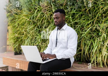 Young African American businessman in formal clothes sitting on bench against hedge and working remotely via netbook Stock Photo