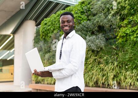 Cheerful young African American male freelancer in white shirt holding netbook while standing near hedge and office building during online work Stock Photo